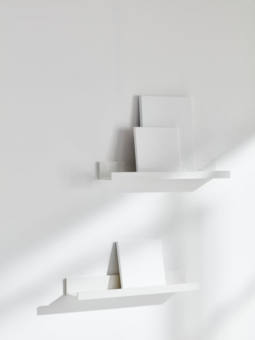 Une pièce aux murs blancs; au mur sont fixées deux tablettes pour photos blanches sur lesquelles reposent des toiles blanches vierges.