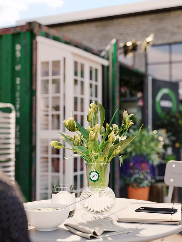A PÅDRAG vase with flowers stands on a grey SUNDSÖ table. There is a napkin, a bowl and a notebook and phone too.