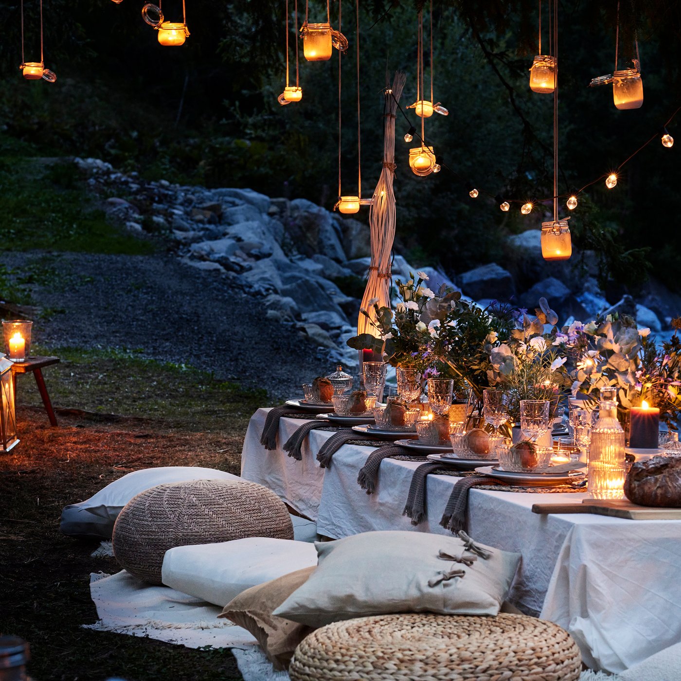 Im Freien neben einem Bach steht ein langer Tisch, gedeckt mit Tischdecke, Geschirr und vielen Blumen. Darum sind verschiedene Sitzkissen und Poufs angeordnet und darüber hängen in der Dämmerung leuchtende Laternen und Lichterketten.