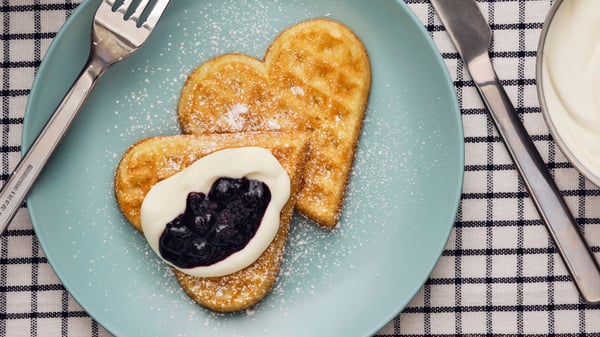 Two heart-shaped waffles with jam, whipped cream and powdered sugar on top are placed on a blue plate.
