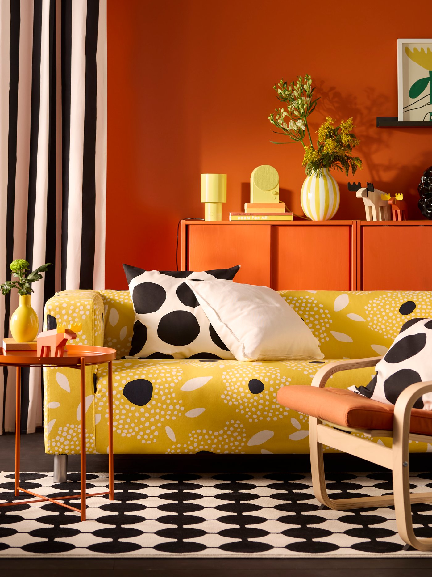 A yellow and white floral-patterned KLIPPAN cover for 2-seat sofa on a sofa in a vivid living room with an orange back wall.