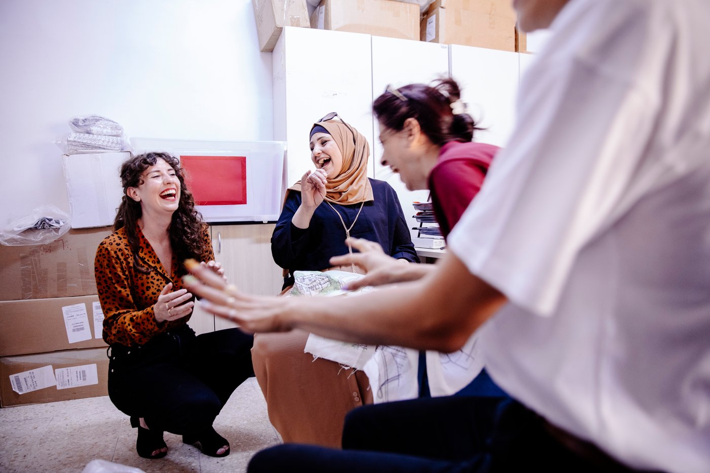 Trzy kobiety siedzące przy stole, jedna kucjająca na podłodze i śmiejące się. 