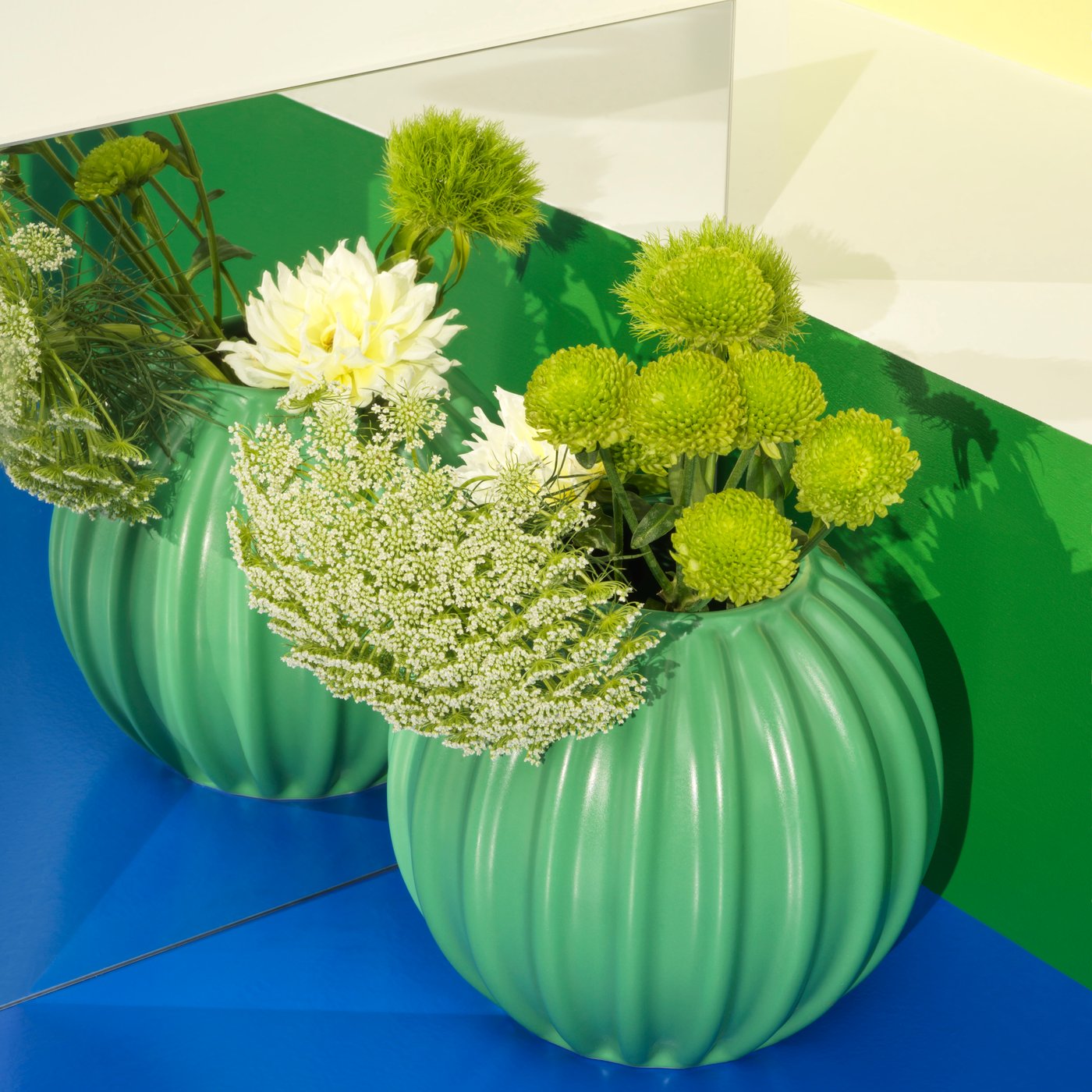 A green SKOGSTUNDRA vase stands on a blue floor before a mirror. Inside the vase are different green flowers.