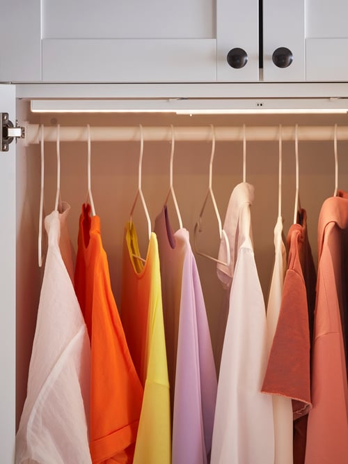 A wardrobe interior has a lighting strip above a clothes rail with colourful shirts on white, indoor/outdoor STAJLIG hangers.