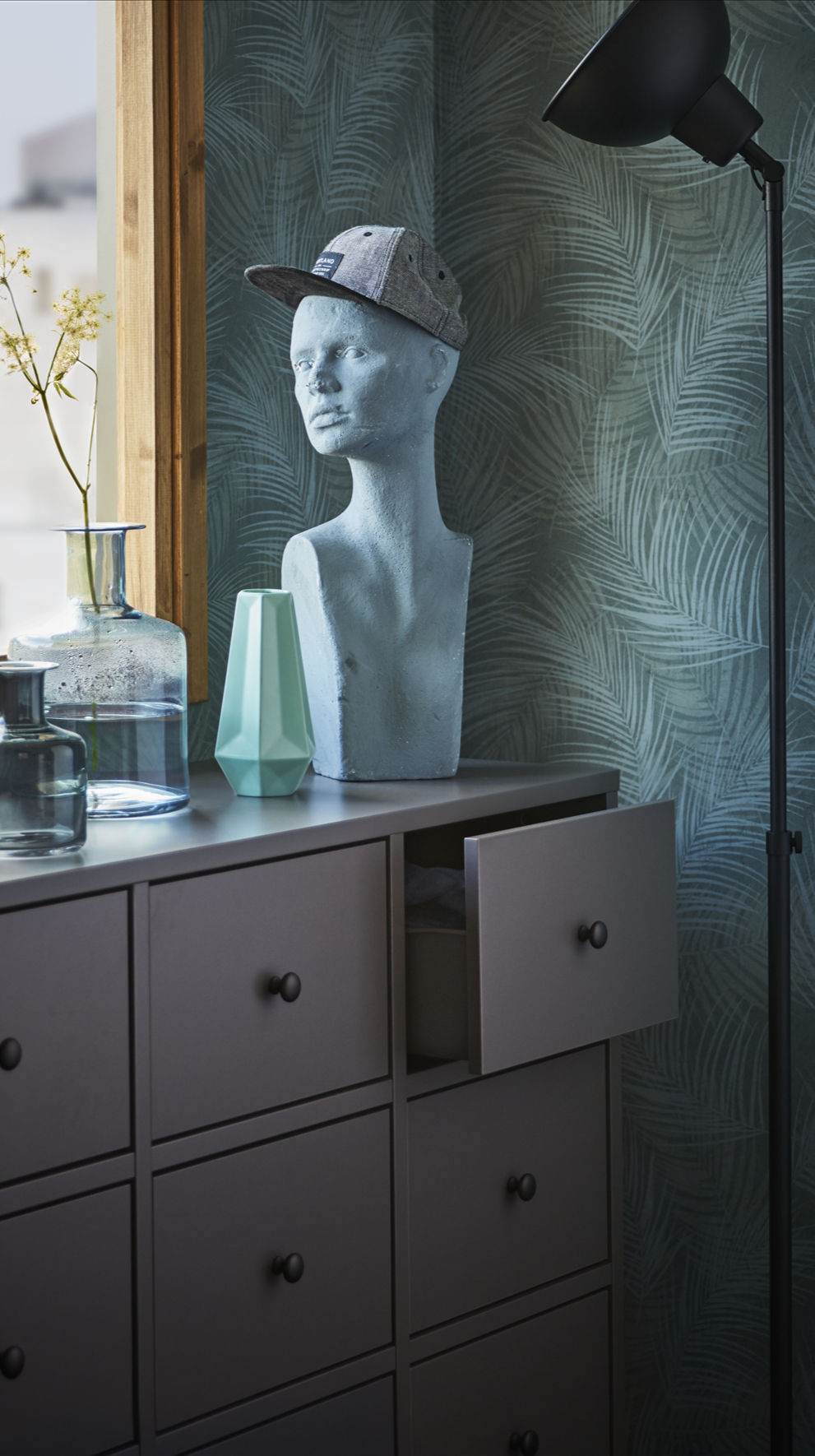 O comodă BRYGGJA gri închis, într-o cameră cu aer primăvăratic. Pe aceasta sunt așezate plante în ghivece, o statuie și alte decorațiuni.