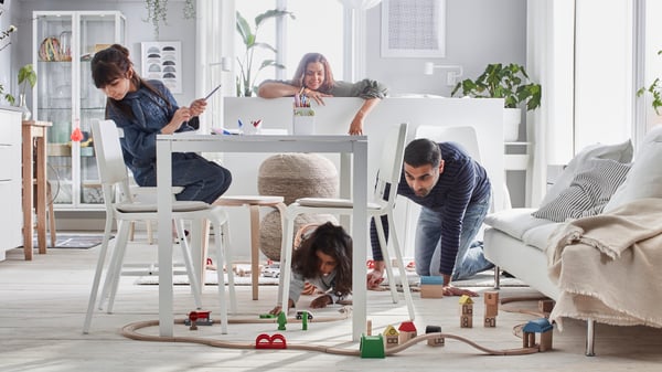 Un adulte à quatre pattes joue avec un enfant sous la table de la salle à manger. Un autre adulte à l’arrière-plan.