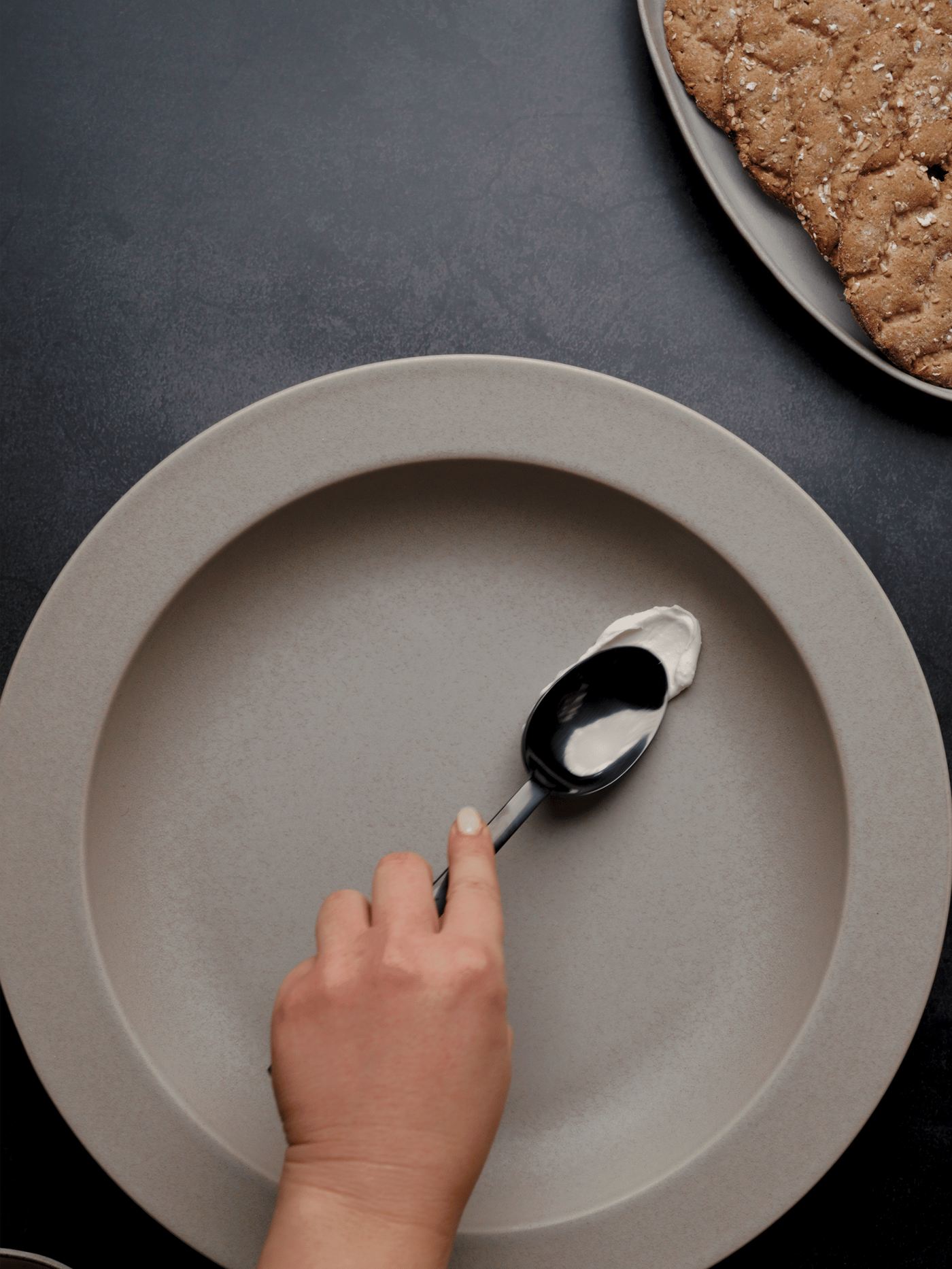 Eine Hand mit einem Löffel nimmt Sahne von einem grauen Teller, im Hintergrund ist ein Keks auf einem Serviertablett zu sehen.