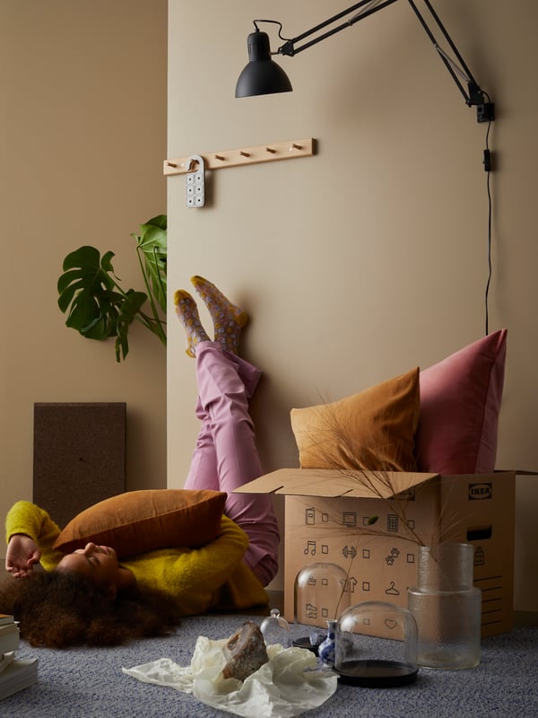 A young woman is laying on the floor. Next to her, stands an IKEA moving box with two cushions in it.
