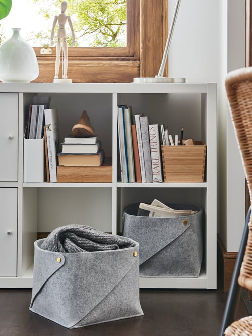 Two grey PUDDA baskets. One is on the floor in front of a KALLAX shelf which has the second one on the lowest shelf.