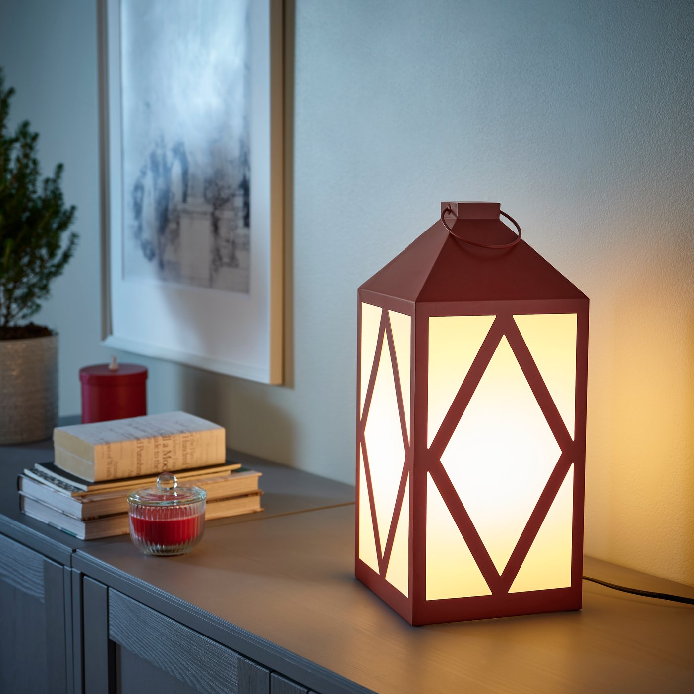 Dekorativ lykta med ljus, böcker, röda ljus i glas och tavla på väggen bakom, placerade på en träyta.