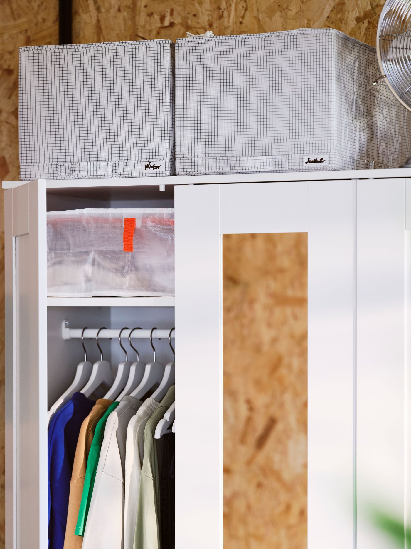 A close-up of a white BRIMNES wardrobe with storage boxes on top. On door open to show hanging shirts in neat order.