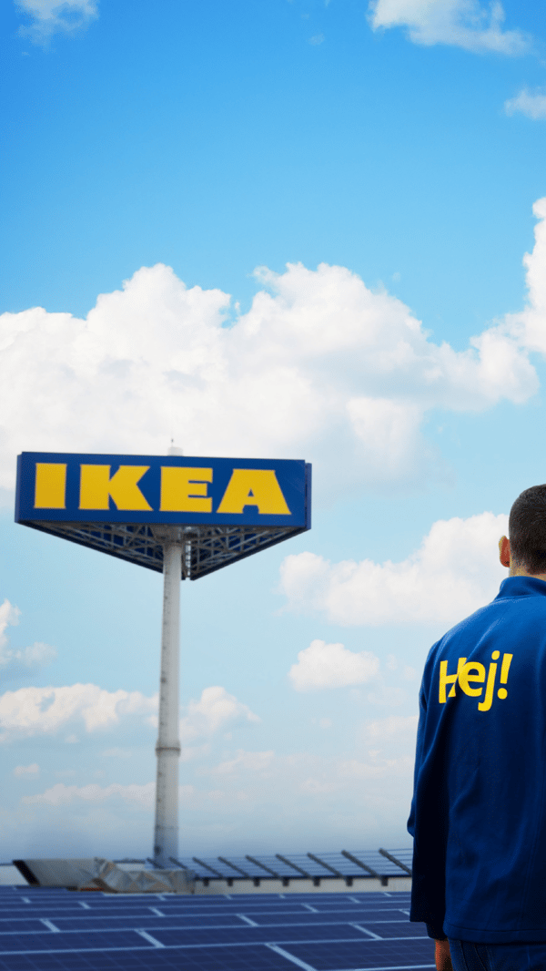 Pracownik IKEA w bluzie z żółtym napisem HEJ na plecach, pozujący na tle słupa z logo IKEA