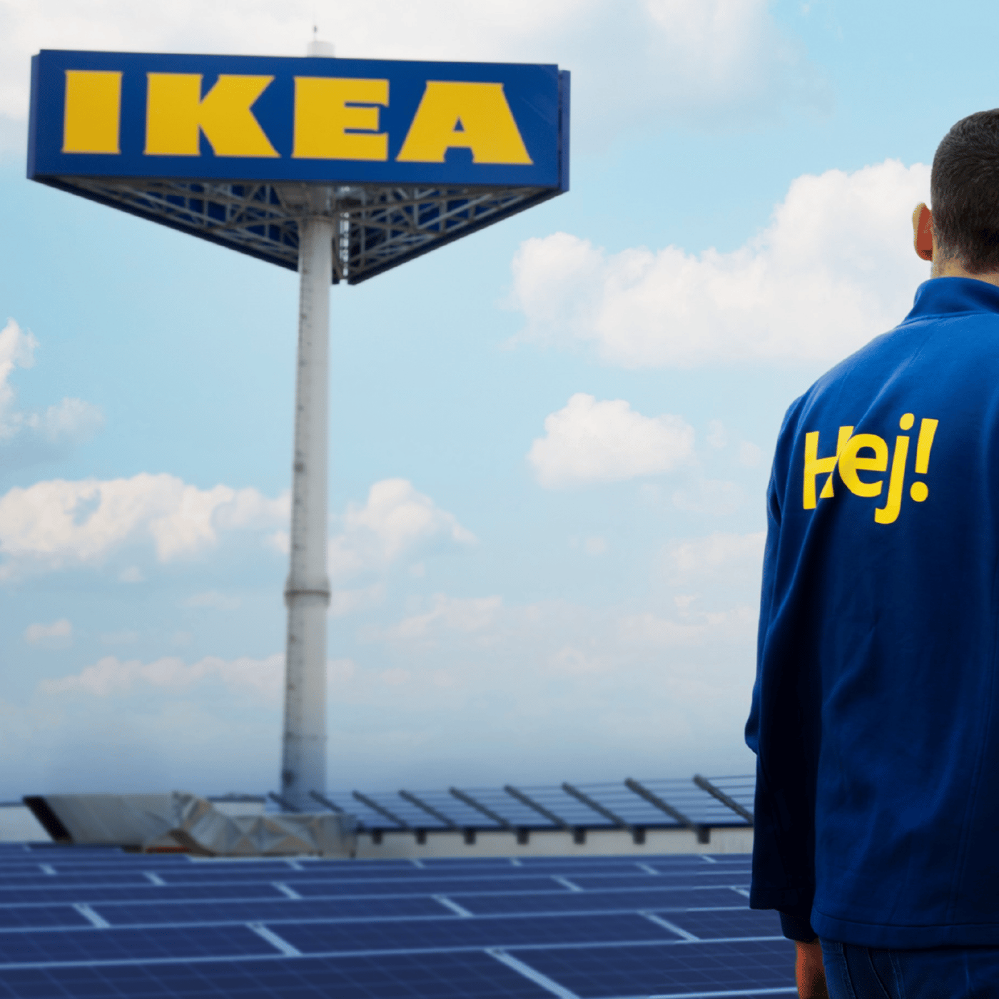 Pracownik IKEA w bluzie z żółtym napisem HEJ na plecach, pozujący na tle pylonu z logo IKEA.