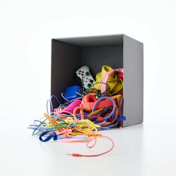 A dark grey DRÖNA box is lying on its side. Inside, we see colourful game consoles, remote controls, headphones and cables.