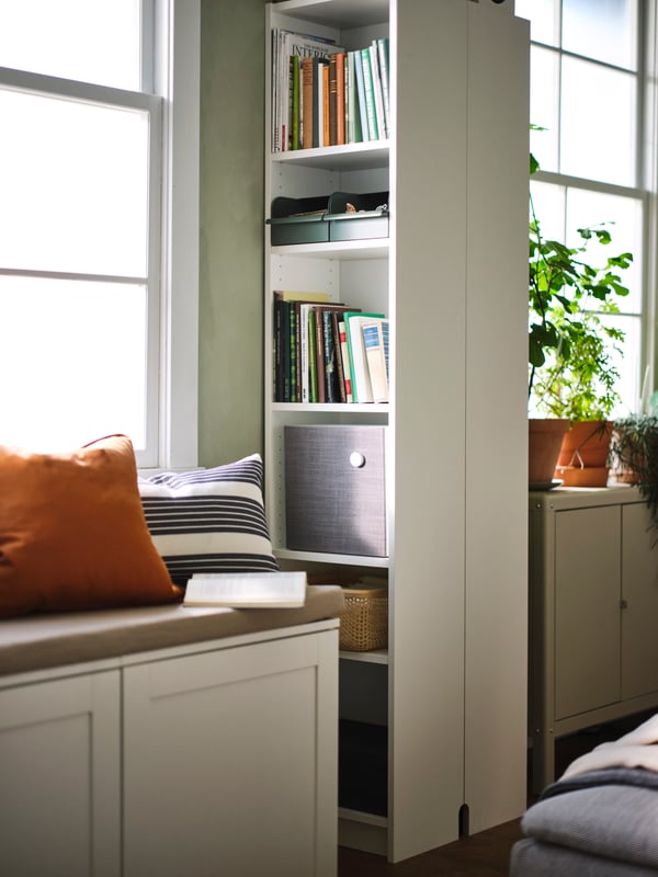 A white PLATSA storage bench in front of two back-to-back white BILLY bookcases, holding books, a speaker and small boxes.