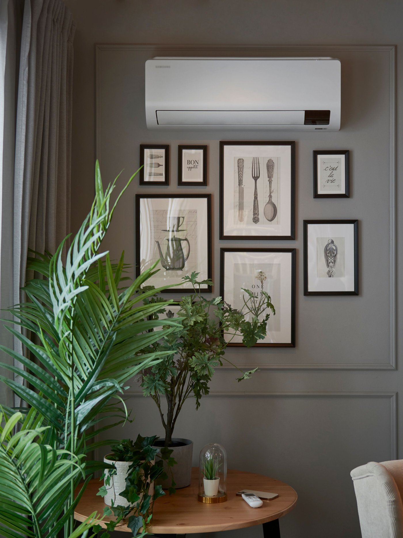 Stylish living room with a wall-mounted air conditioner, botanical artwork, lush green plants, and a round wooden table.