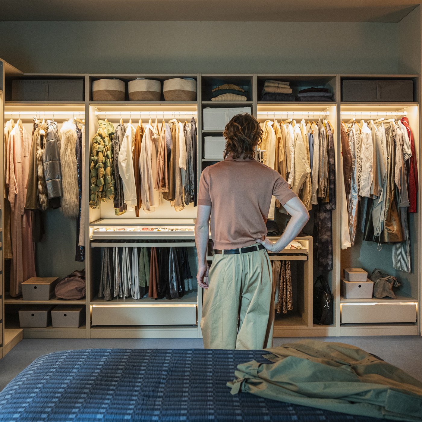 Una persona in piedi di spalle guarda un ampio armadio aperto e illuminato, pieno di vestiti appesi e piegati.