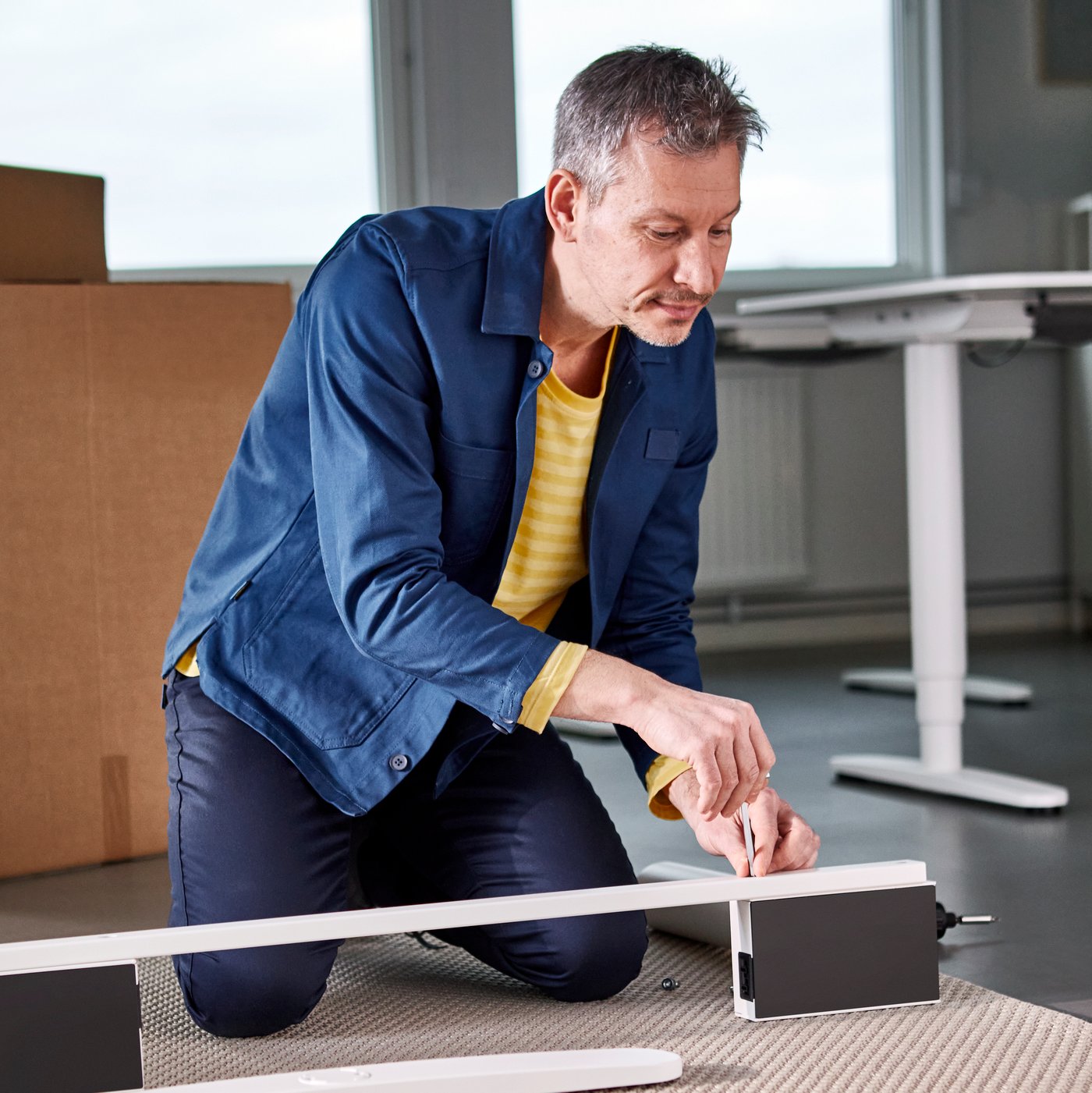 Zaměstnanec IKEA, který montuje kancelářský stůl BEKANT, zatímco klečí na koberci.