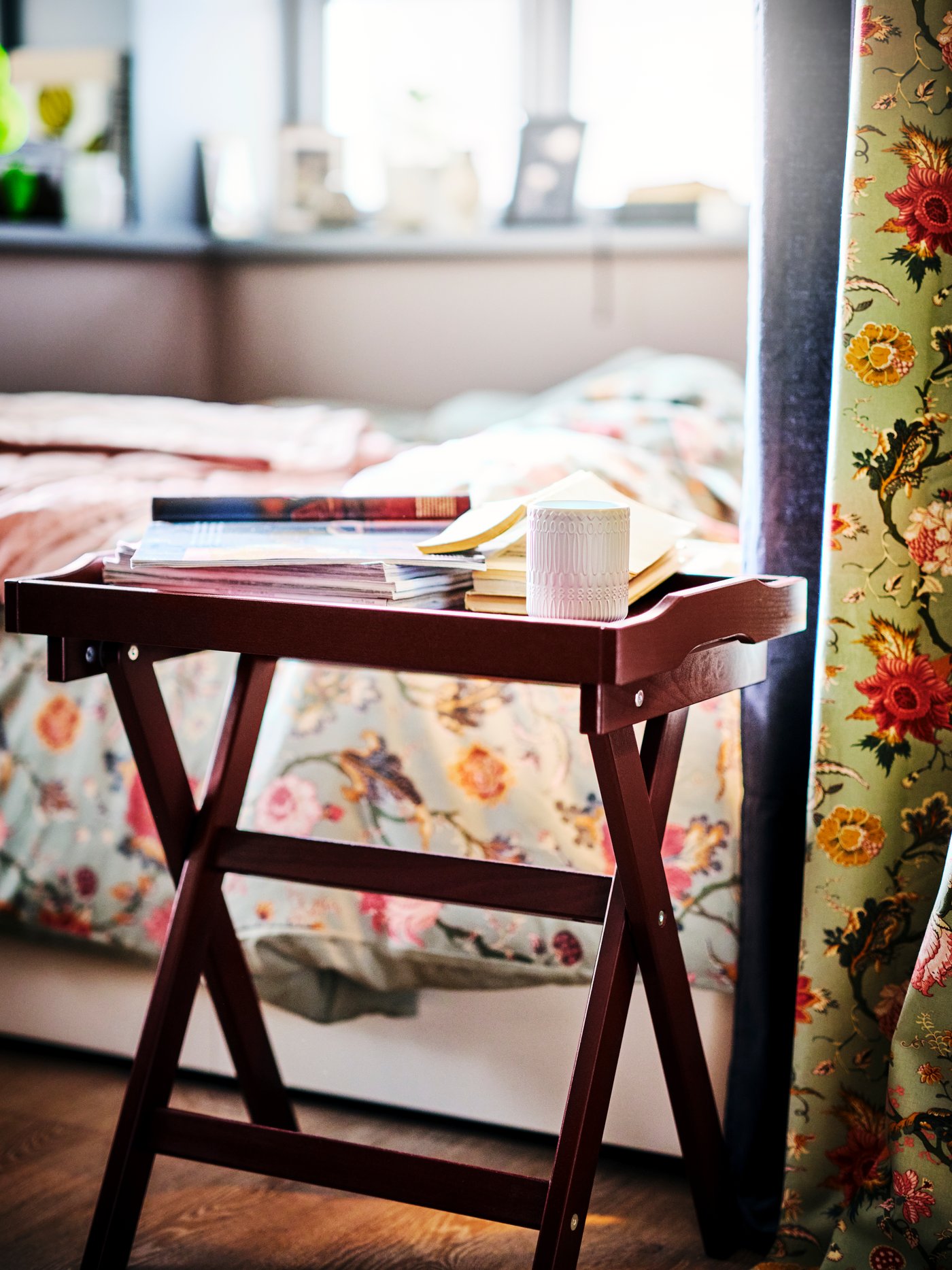 A dark red MARYD tray table with books next to a bed with a floral bedspread with a matching floral curtain to the right.