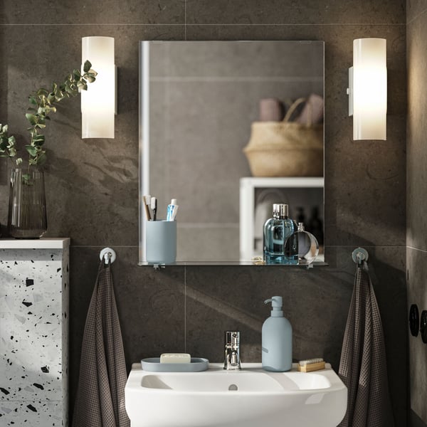A rectangular NYSJÖN mirror with a glass shelf holds toothbrushes and perfume bottles above a white wash-basin and cabinet.