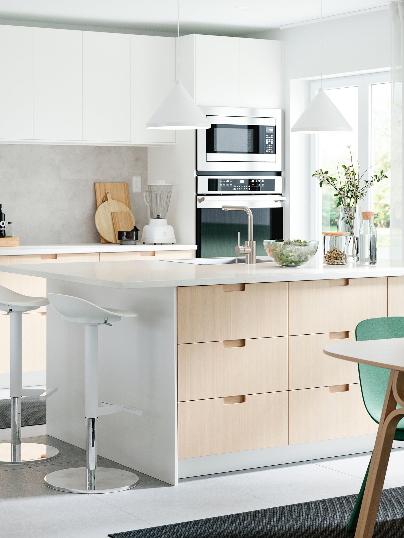 A kitchen with drawer fronts and doors in white/bamboo, a kitchen island, two bar stools, two pendant lamps and green chairs.