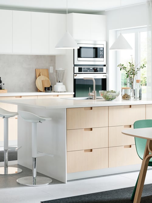 A kitchen with drawer fronts and doors in white and bamboo, a kitchen island, two bar stools, two pendant lamps and green chairs.