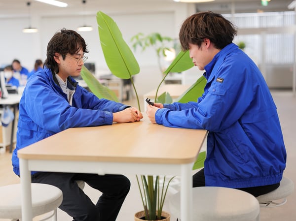 ブルーの制服を着た2人が白いテーブルを挟んで向かい合い、観葉植物のある明るいオフィスで会話している。