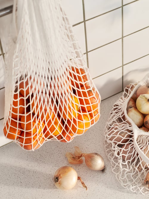 Un coin cuisine dans lequel se trouvent deux sacs en filet, un est suspendu à un comptoir et contient des oranges, tandis que l’autre est rempli d’oignons. 