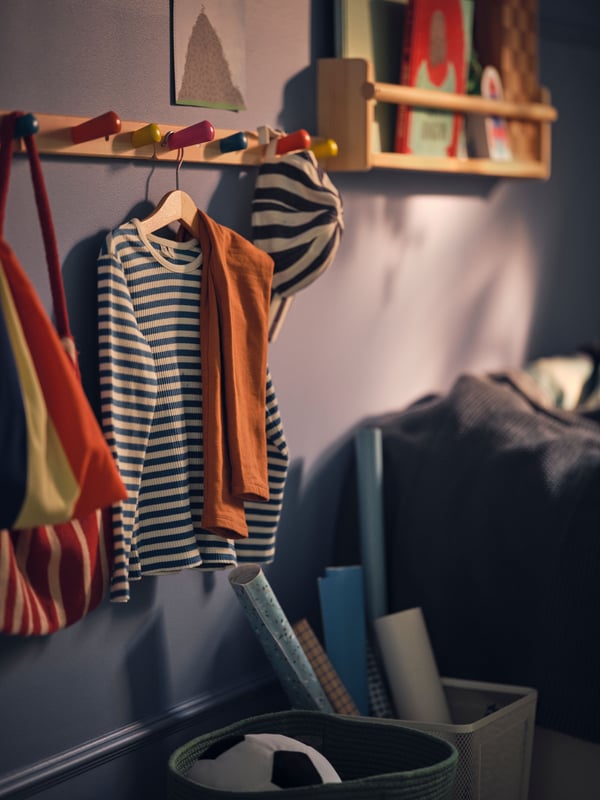 A blue wall with a FLISAT wall storage holding books on it next to a FLISAT knob rack with 4 knobs holding clothes and a hat.