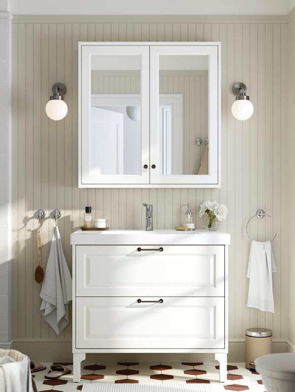 A white TÄNNFORSEN washstand is placed in a white bathroom below a white mirror cabinet with two white lamps on either side.