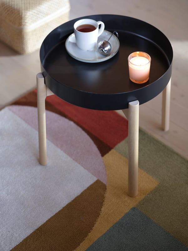 A multicoloured rug with a geometrical pattern, a black YPPERLIG coffee table with birch legs, a cup of tea and a candle.