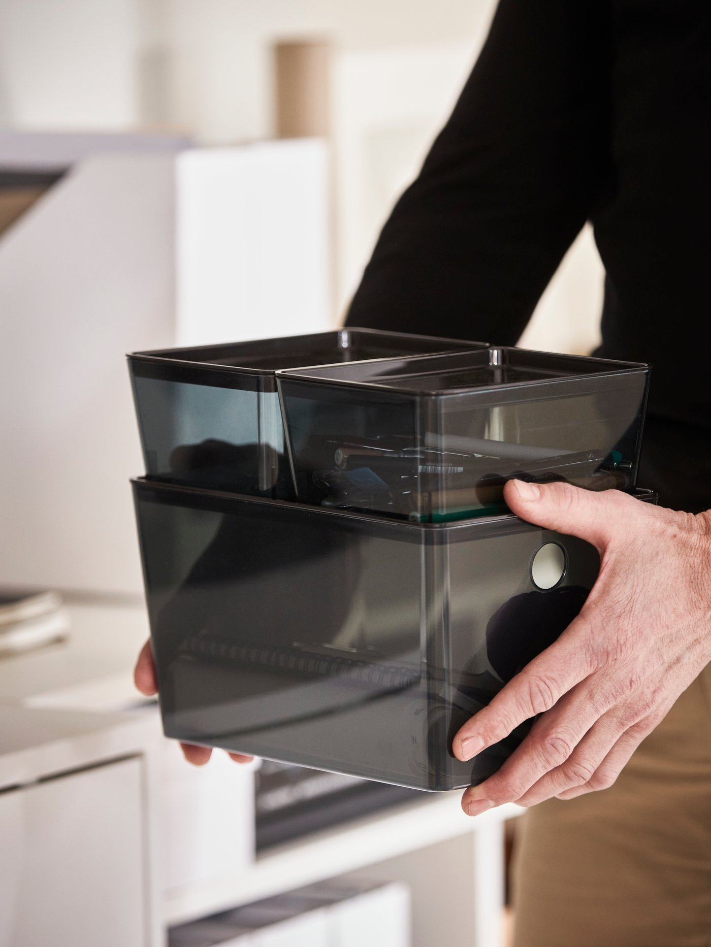 Close-up of a man holding three black KUGGIS storage boxes stacked together. A smaller box shows pens stored inside.