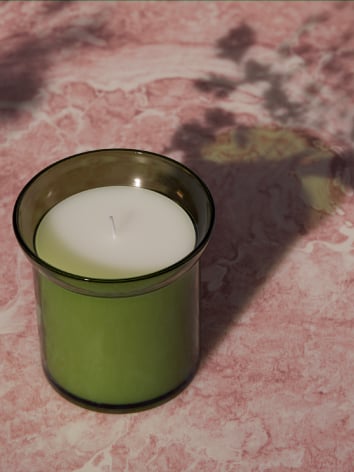 شمعة معطرة HEDERSAM في مرطبان زجاجي أخضر على سطح رخامي وردي، تلقي بظلال على شكل زهرة.