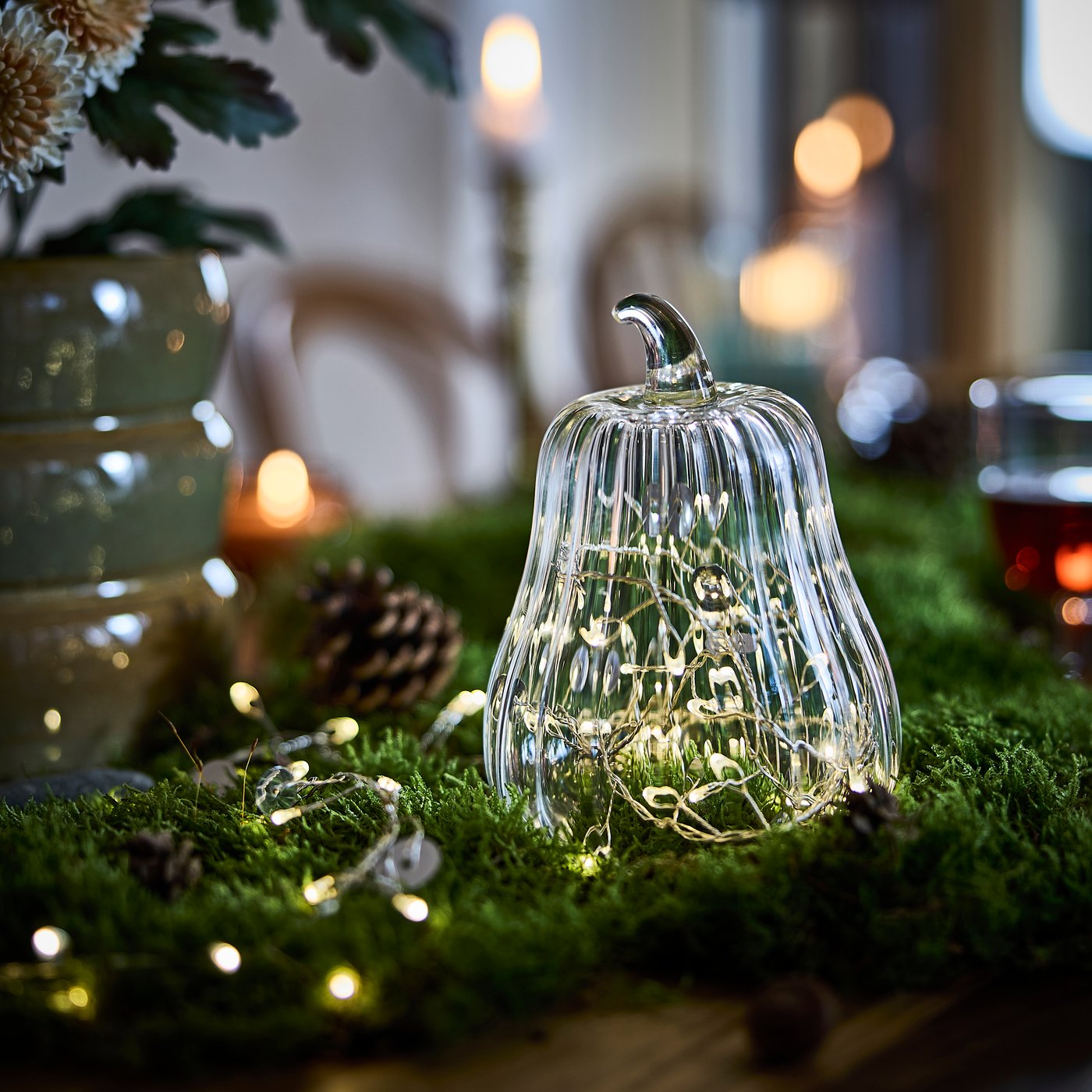 Una campana de vidrio HÖSTAGILLE con forma de calabaza con una guirnalda de luces en su interior; se ha colocado sobre musgo, en una mesa con temática forestal.