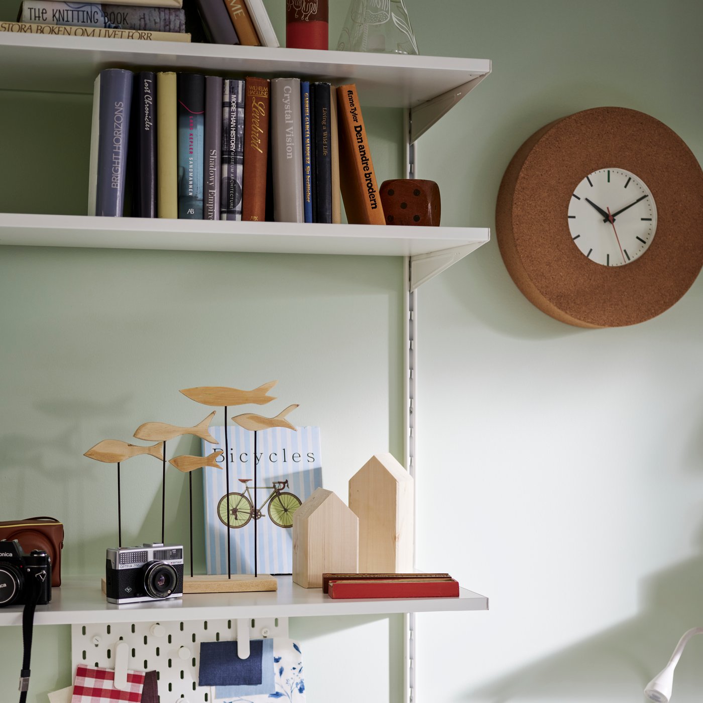 A SVARTGÖK wall clock mounted on a light green wall beside white shelves holding books and small decorative items.