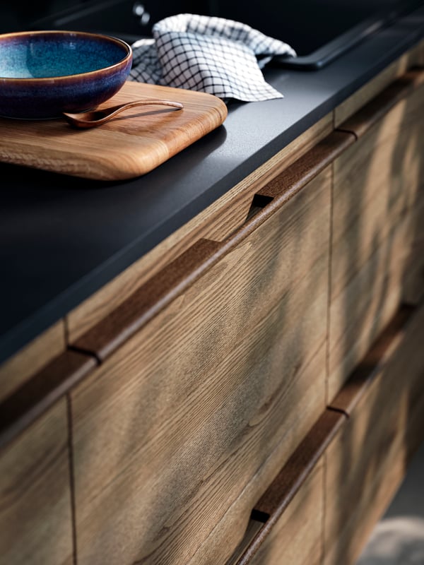 Des façades de tiroirs en placage de frêne marron foncé STENSTA, un comptoir noir élégant, une planche à découper en bois et un bol en céramique sur le dessus.
