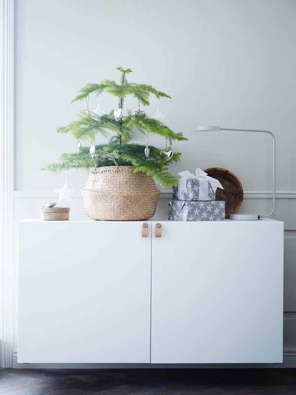 Due mobili con ante BESTÅ su cui è appoggiate un cestino FLÅDIS con dentro una pianta con decorazioni. Accanto al cestino due pacchetti regalo e una confezione di legno. 