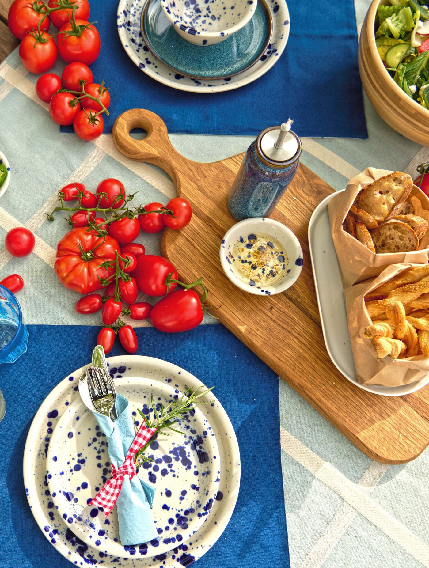 Picnic table with tomatoes, bread, salad and ceramic dishes on a blue-and-white tablecloth.