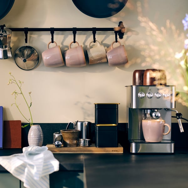 A kitchen with a worktop area holding various kitchen appliances and FÄRGKLAR cups hanging from a wall rack above.