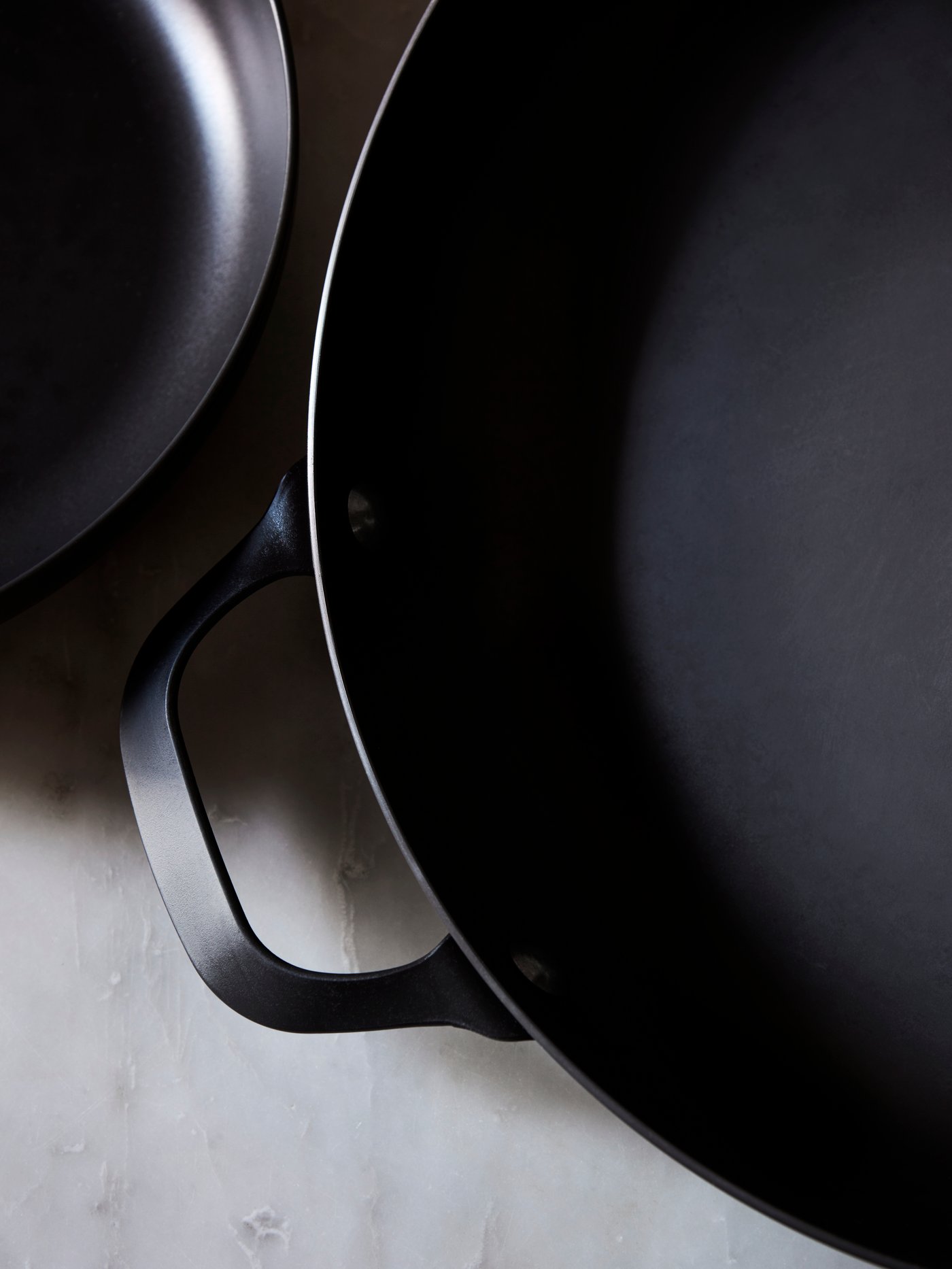 A close-up of the black carbon steel VARDAGEN frying pan with a handle placed on a grey marble countertop.