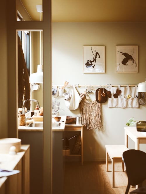 Un cărucior RÖNNINGE amplasat într-o bucătărie. Alături, o masă extensibilă cu o bancă și, deasupra, o șină cu decorațiuni.