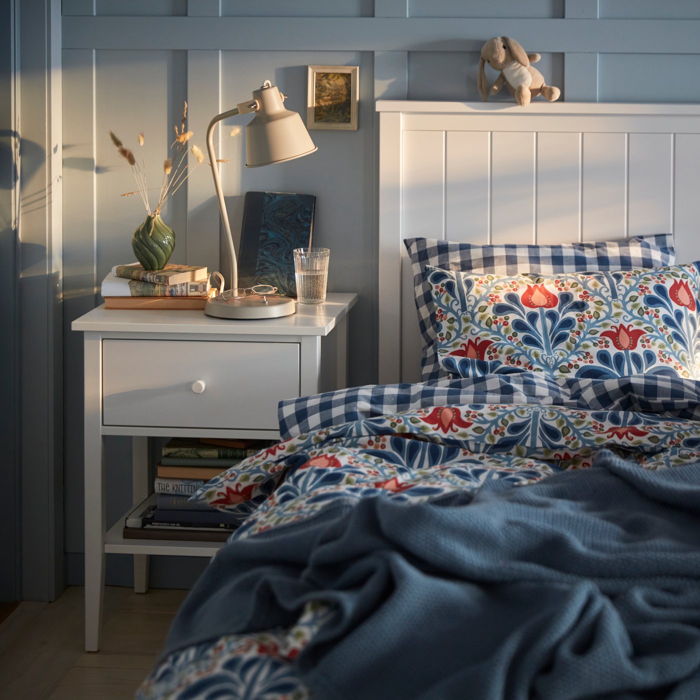 A white bed with a floral multicoloured SÄTERVIDE duvet cover set next to a white bedside table with a lamp.