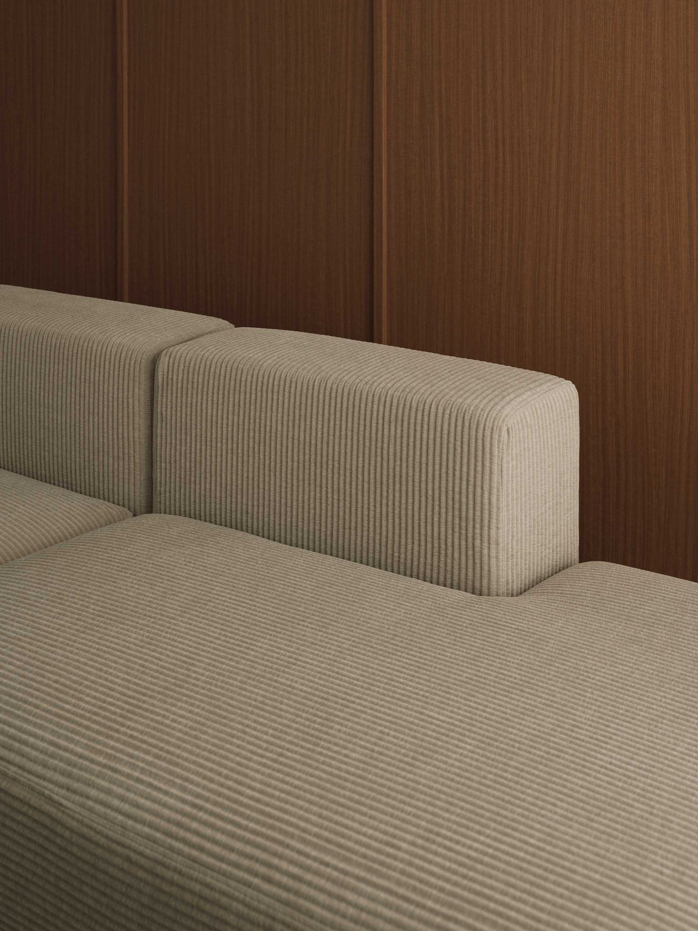 A close-up of a wonderfully comfortable light beige modular sofa, featuring a lovely ribbed upholstery that adds a touch of texture and cosiness to your living space. The sofa sits against a warm brown paneled wall, creating a harmonious and inviting backdrop. This image highlights the inviting details of the sofa, perfect for relaxing and unwinding after a long day.