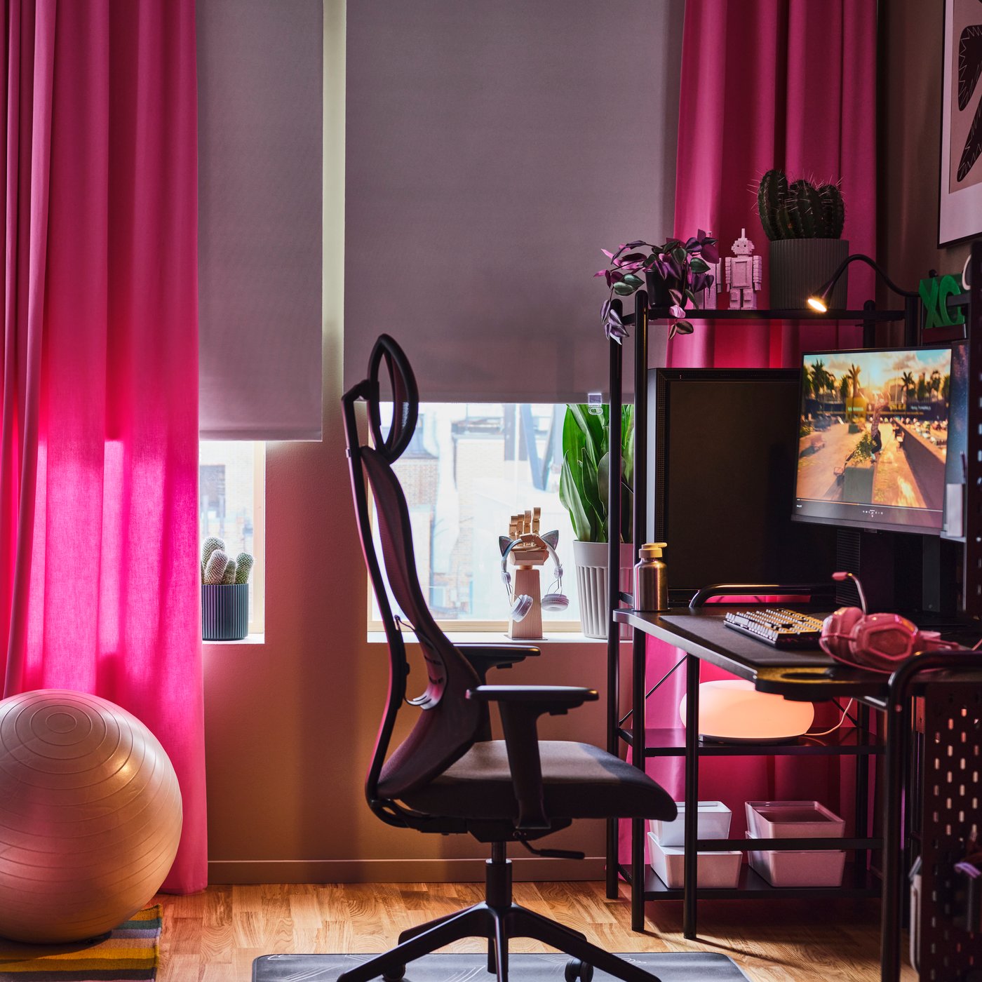 Una oficina en casa con un escritorio gamer FRÖSKELÅS negro con una computadora. En el alféizar de una ventana con cortinas rosas hay varias plantas.