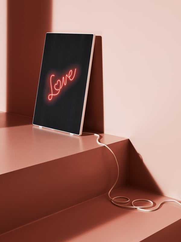 Ein SYMFONISK Rahmen-Lautsprecher steht auf einer rosa Treppe, darauf leuchtet in Neon-Schrift das Wort ‚Love‘ – das Kabel ist locker aufgerollt