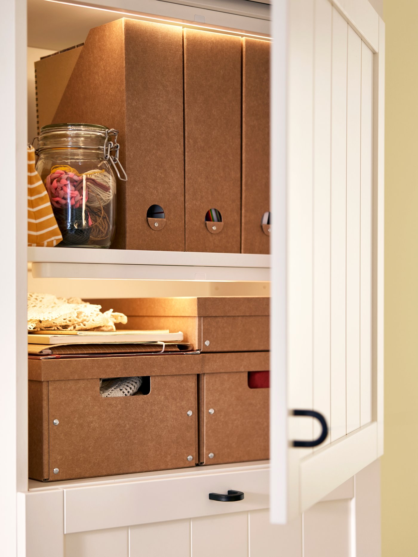 Pojemniki GÄRDESGÅRD z pokrywkami i segregatory ustawione na półkach białej szafki BESTÅ z uchylonymi drzwiami PIPMAKARE.