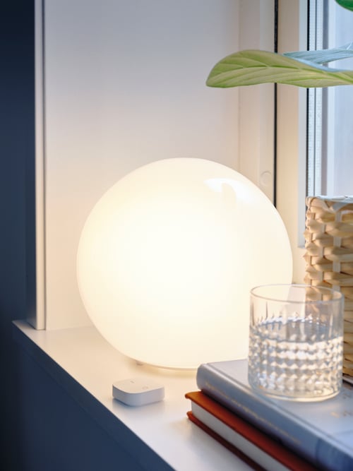 Un dormitor modern, dotat cu o veioză, un dimmer wireless, o oală de plante și un pahar de whisky, toate așezate pe un pervaz.