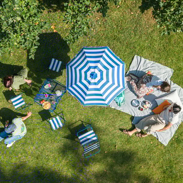 Friends have a barbecue in the park, relaxing on the grass with a STRANDÖN table, chairs, stools, and parasol.
