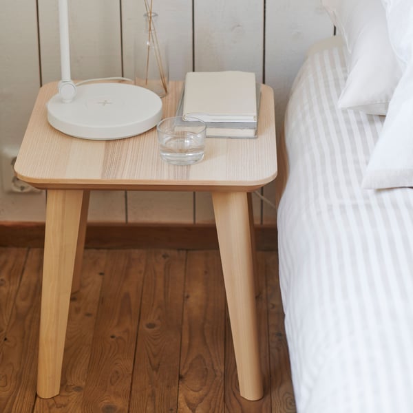 LISABO side table next to a bed frame with NATTJASMIN duvet covers. A NYMANE table lamp, IKEA 365+ glass and books are placed on the table. 