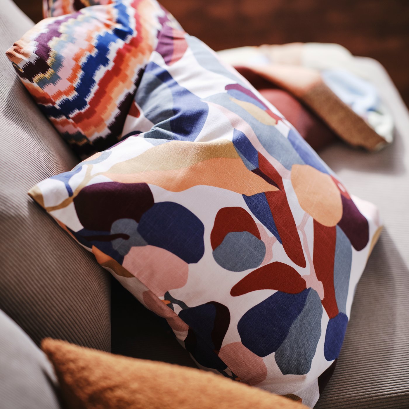 A close-up of a multicoloured VIDEOXBÄR cushion cover with abstract floral patterns on a beige sofa.